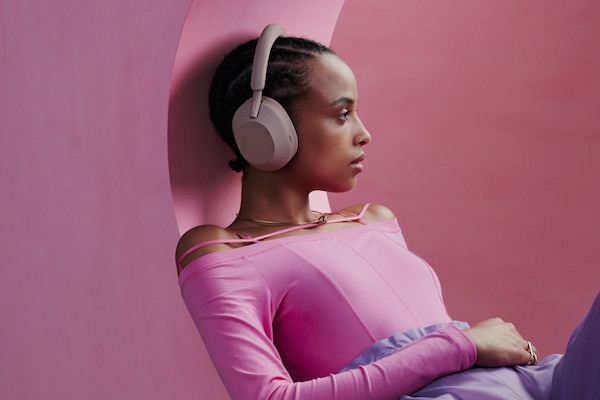 A promotional shot of a girl leaning against a pink wall wearing her WH-1000XM5 wireless headphones.